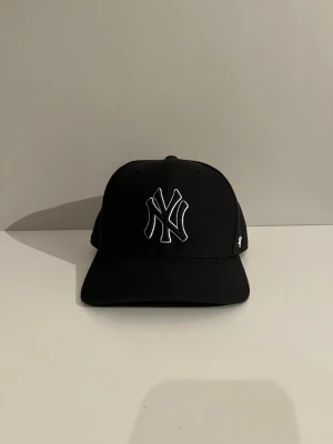 Svart New York Yankees keps från 47 - Snygg svart keps med böjd skärm från 47, prydd med New York Yankees-logga i vitt framtill. Klassisk passform och broderad logotyp på sidan. Tillverkad i ett slitstarkt tyg som ger en clean och sportig look.