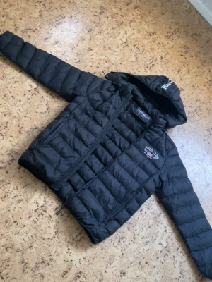 Svart dunjacka från Polo Club - Snygg svart dunjacka från Polo Club med huva och dragkedja. Jackan har broderad logga på bröstet och text på huvan. Lättviktig och quiltad design som håller dig varm under kalla dagar. Perfekt för dig som vill ha en stilren och sportig look.