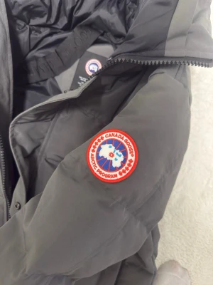 Mörkgrå dunjacka från Canada Goose - Säljer en mörkgrå dunjacka från Canada Goose med klassisk patch på ärmen. Jackan har huva, dragkedja och tryckknappar framtill samt två stora fickor. Perfekt för kalla vinterdagar och riktigt varm tack vare dunfyllningenoch vattentätt 