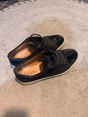 Svarta sneakers från Lanvin - Säljer nu mina snygga navy sneakers från Lanvin då jag är sugen på något annat. Ovandel i mocka och lackad tå. Skorna har vita sulor och klassisk snörning.  Storlek 8, passar mig som har 43/44 bra. Inga större defekter, litet märke fram på högra skon,  går att se på vissa ställen mockan inte är i nyskick. Låda, snören m.m finns dock inget kvitto. Hör av dig vid fundering!