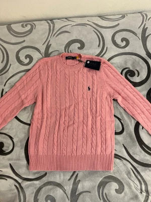 Rosa kabelstickad tröja Polo Ralph Lauren - Säljer en rosa kabelstickad tröja från Polo Ralph Lauren. Tröjan har rund hals, långa ärmar och den klassiska broderade loggan på bröstet. Perfekt för dig som vill ha en stilren och tidlös look med en mjuk känsla.