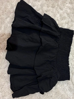 Svart volangkjol med resårmidja - Kort svart kjol med bred resår i midjan och flera volanglager som ger en luftig och trendig look. Perfekt att matcha med en crop top eller stickad tröja. Kjolen har en avslappnad passform och är tillverkad i ett mjukt tyg.