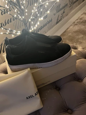 Svarta sneakers från Axel Arigato - Stilrena svarta sneakers från Axel Arigato med vit sula och diskret logga på sidan. Skorna har en klassisk låg profil och är tillverkade i mocka och läder för en lyxig känsla. Perfekta för dig som gillar minimalistisk och modern design. Storlek 40. Går att fräscha till lite väldigt simpelt. PRIS kan diskuteras
