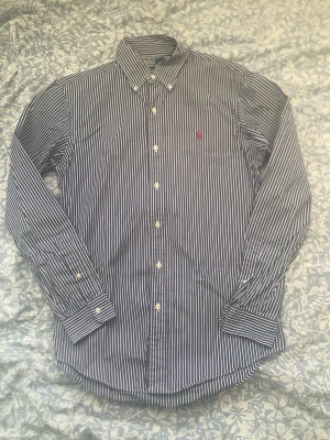 Randig skjorta från Ralph Lauren - Klassisk blå och vit randig skjorta från Ralph Lauren med lång ärm och knappar hela vägen. Skjortan har en broderad röd logga på bröstet och är tillverkad i bomull. Perfekt för dig som gillar stilrena och tidlösa plagg. Väldigt bra skick men storlekslappen är dock bortklippt.                          Grisch, polo Ralph lauren