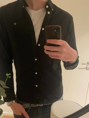 Svart skjorta från Morris Stockholm - Säljer en svart skjorta från Morris Stockholm med vita knappar och klassisk krage. Skjortan har en liten broderad logga på bröstet och är långärmad. Perfekt för dig som gillar stilrena plagg med snygga detaljer.