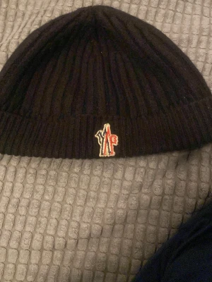 Svart Moncler ribbstickad mössa - Svart ribbstickad mössa från Moncler med klassisk uppvikt kant och broderad logotyp framtill i rött, vitt och blått. Perfekt accessoar för att hålla värmen och ge en stilren touch till din outfit.