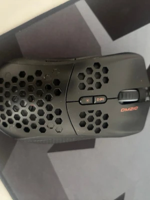 Svart gamingmus DM210 med hål - Säljer en svart gamingmus DM210 med honeycomb-design för lättare vikt. Musen har DPI-knapp, scrollhjul och två sidoknappar. Tillverkad i plast med mattsvart finish och orange DM210-logga. Perfekt för gaming och snabba rörelser.