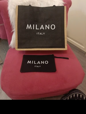 Svart shoppingväska Milano Italy - Snygg svart shoppingväska från Milano Italy med beige kanter och vit text. Tillverkad i grovt tyg, Medföljer en matchande mindre necessär med dragkedja. Perfekt för dig som vill ha en trendig och rymlig väska till vardagen.