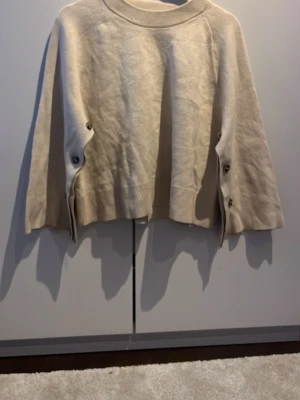 Beige stickad tröja med knappar - Säljer en beige stickad tröja med vida ärmar och snygga knappar längs sidorna. Tröjan har rund halsringning och ribbade muddar nedtill. Perfekt för dig som gillar en avslappnad och trendig stil.
