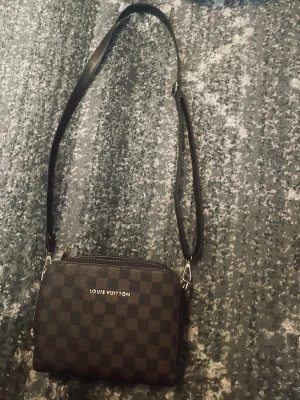 Louis Vuitton axelväska brun - Snygg axelväska från Louis Vuitton i klassiskt brunt damier-mönster. Väskan har justerbar svart axelrem och silverfärgade detaljer. Perfekt storlek för det viktigaste och stängs med dragkedja. Ikonisk logga framtill.