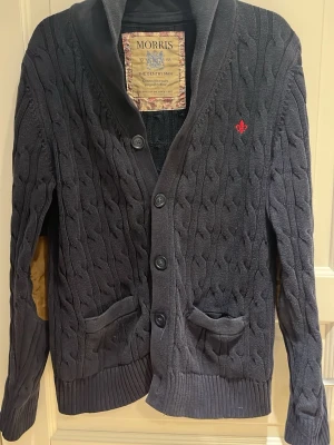 Morris kofta mörkblå  - Snygg och väldigt sällsynt mörkblå kabelstickad kofta från Morris med tre knappar framtill, två fickor och ribbade muddar. Koftan har en liten röd brodyr på bröstet och klassisk krage. Perfekt för lager på lager och stilrena outfits.