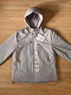 M jacka - Snygg ljusgrå vindbreaker med M loggan på axeln. Jackan har text längs dragkedjan, två fickor med dragkedja och justerbar nederkant. Tillverkad i lätt, vattenavvisande material. Perfekt för blåsiga dagar. Jackan är helt ny, oanvänd. Kan passa S också. Priset är diskuterbart.