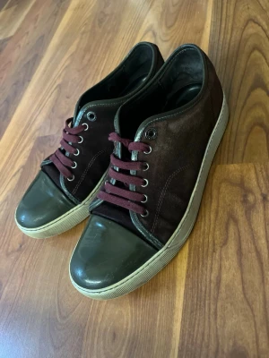 Lanvins - Snygga sneakers i mörkbrunt mockaliknande material med vinröda snören och tåparti i blank mörkgrön plast. Yttersulan är vit och ger en klassisk look. Perfekta för dig som gillar en stilren men unik touch på dina skor.