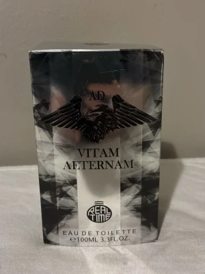 Ad Vitam Aeternam Eau de Toilette - Ad Vitam Aeternam Eau de Toilette från Real Time i en cool, transparent glasflaska med silverfärgat lock och svart logga. Doften kommer i en 100 ml flaska och har en modern, edgy design med vingar och geometriska mönster på förpackningen.