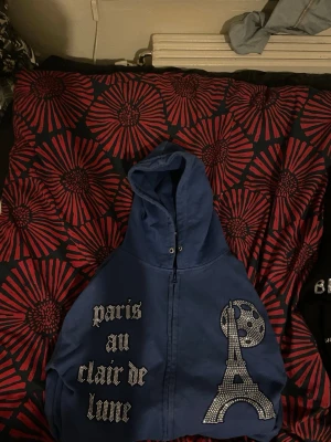 Blå bling lucid  hoodie med Paris-motiv - Säljer en blå hoodie med dragkedja och huva. På framsidan finns texten 'paris au clair de lune' i glittriga stenar samt ett motiv av Eiffeltornet och månen i samma stil. Hoodien har en avslappnad passform och är perfekt för dig som gillar statement-plagg.