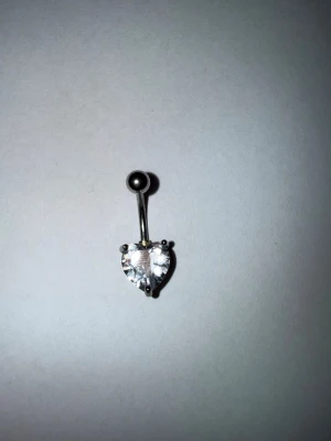 Navelpiercing med hjärtformad sten - Snygg navelpiercing i silverfärgad metall med en glittrande, hjärtformad klar sten. Perfekt accessoar för att ge din look lite extra bling. Enkel och stilren design som passar till många olika stilar.