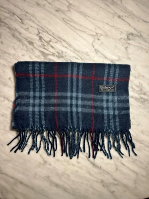 Mörkblå rutig halsduk från Burberry - Klassisk halsduk i mörkblått med rutigt mönster i ljusblått och rött från Burberry. Halsduken har fransar i kanterna och en vävd Burberry-etikett. Tillverkad i mjukt material som känns lyxigt och stilrent.