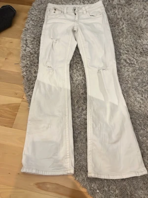 Vita bootcut jeans med slitningar - Säljer ett par vita bootcut jeans med slitningar vid knäna. Jeansen har fem fickor, låg midja och vida ben. Perfekta för dig som gillar en avslappnad och trendig stil.