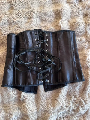Svart satin korsett med snörning - Snygg svart korsett i satin med klassisk snörning framtill och hakar för stängning baktill. Modellen har markerade sömmar och ger en riktigt cool siluett. Perfekt för att lyfta vilken outfit som helst.