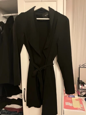 Svart tunn kappa från Vila med knytskärp - Stilren svart kappa från Vila med lång ärm och knytskärp i midjan. Kappan har en öppen front med bred krage och ett mjukt, följsamt tyg som ger en snygg siluett. Perfekt för dig som vill ha en enkel men trendig look.