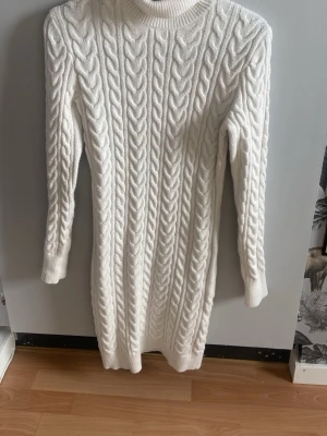 Vit stickad klänning från FB Sister - Mysig vit stickad klänning från FB Sister med kabelstickat mönster och lång ärm. Klänningen har en rund halsringning och är perfekt för kyligare dagar. Passformen är normal och materialet känns mjukt och varmt.