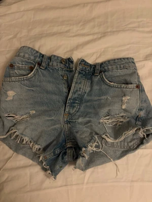 Ljusblåa jeansshorts - Säljer ett par ljusblåa high waist jeansshorts med slitna detaljer och fransig kant. Klassisk femficksmodell med knappgylf och bälteshällor. Perfekta för sommaren och ger en avslappnad vibe. skriv om ni vill ha mer bilder 