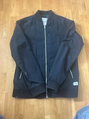 Svart bomberjacka från Jack & Jones - Stilren svart bomberjacka från Jack & Jones med silverfärgad dragkedja framtill och två fickor med dragkedja. Jackan har ribbade muddar vid ärmslut och nederkant samt en diskret logolapp vid ena fickan. Fodret är mörkgrönt och materialet känns lätt och smidigt.