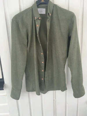 Olivgrön skjorta  - Stilren olivgrön skjorta med klassisk krage och ljusa knappar. Skjortan har lång ärm och är tillverkad i ett mjukt bomullstyg som känns skönt mot huden. Perfekt för dig som gillar en enkel men snygg look.