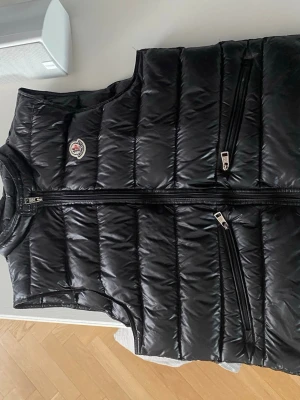 Svart dunväst från Moncler - Snygg svart dunväst från Moncler med glansig finish och klassisk logga på bröstet. Västen har två snedställda dragkedjefickor och hög krage med tryckknapp. Perfekt för lager-på-lager och riktigt trendig look.