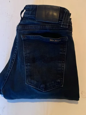 Mörkblå jeans från Nudie Jeans - Snygga mörkblå jeans från Nudie Jeans med klassisk femficksmodell och vågig söm på bakfickan. Jeansen har en läderpatch i midjan och är tillverkade i slitstark denim. Perfekta för dig som gillar en tidlös och clean look.