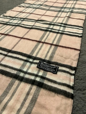 Beige rutig halsduk från Burberrys - Klassisk beige halsduk från Burberrys i 100% lammull med ikoniskt rutmönster i svart, vitt och rött. Halsduken har fransar i ändarna och en vävd etikett. Perfekt accessoar för att lyfta din outfit.