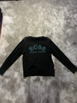 Svart sweatshirt från Hugo Boss - Svart sweatshirt från Hugo Boss med blått BOSS-tryck på bröstet. Tröjan har rund halsringning, långa ärmar och ribbade muddar. Perfekt för en stilren och avslappnad look. Är storlek l men sitter mer som s-m