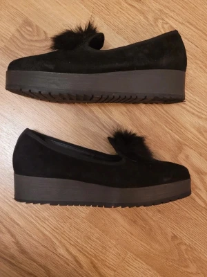 Svarta loafers med platåsula och pälsdetalj - Säljer ett par svarta loafers med hög platåsula och fluffig pälsdetalj framtill. Skorna är i mockaliknande material och har en chunky, modern look. Perfekta för dig som vill ha något extra till outfiten. Snygga och trendiga skor som sticker ut.