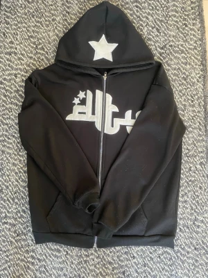 Svart hoodie med stjärntryck - Säljer en svart hoodie med dragkedja och stor huva. På bröstet finns ett stort tryck i vitt med stjärnor och text, samt en stor vit stjärna på huvan. Hoodien har fickor framtill och ribbade muddar vid ärmar och nederkant. Perfekt för en chill och street-inspirerad look.