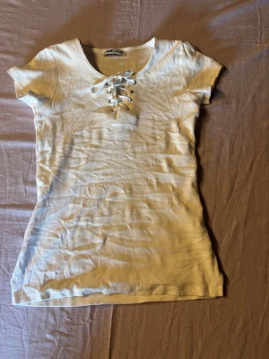 Vit ribbad topp med snörning - Kortärmad vit topp med ribbad struktur och snygg snörning framtill vid urringningen. Figurnära passform och mjukt material som känns skönt mot huden. Perfekt till jeans eller kjol för en trendig look.