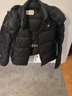 Svart pufferjacka Calvin Klein  - Snygg svart pufferjacka från Calvin Klein Jeans med stor logga på insidan och mindre CK-detalj på bröstet. Jackan har hög krage, dragkedja och tryckknappar samt vadderad design som håller dig varm under kalla dagar. Perfekt för vintern och riktigt trendig. Mycket bra skick varsamt använd 