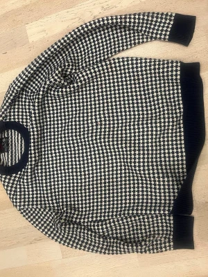 Stickad tröja med svartvitt mönster - Snygg stickad tröja från COS med svart och vitt rutmönster. Tröjan har rund halsringning, ribbade muddar och nederkant i svart. Perfekt för dig som gillar en stilren och modern look.
