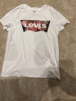 Vit Levi's t-shirt med blommigt tryck - Snygg vit t-shirt från Levi's med klassiskt logotryck på bröstet, där bakgrunden är fylld med rosa blommor. T-shirten har rund hals och korta ärmar. Perfekt för dig som vill ha en enkel men ändå unik look.