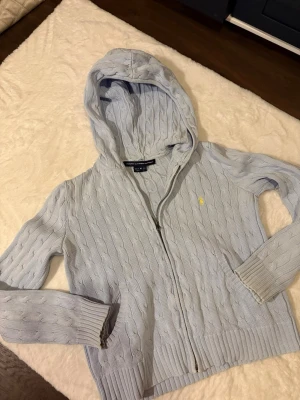 Ljusblå kabelstickad hoodie Polo Ralph Lauren - Ljusblå kabelstickad hoodie från Polo Ralph Lauren med dragkedja framtill och fickor på sidorna. Tröjan har ribbade muddar och en liten gul broderad logga på bröstet. Perfekt för en avslappnad och stilren look.