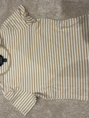 Randig beige/vit t-shirt Lauren Ralph Lauren - Snygg randig t-shirt från Lauren Ralph Lauren i beige och vitt. Klassisk rund halsringning och korta ärmar. Tillverkad i mjuk bomull som känns skön mot huden. Perfekt för en avslappnad och stilren look. Ny utan prislapp, pris kan diskuteras vid snabb affär