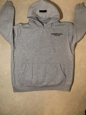  Essentials Fear of God hoodie - Snygg grå hoodie från Essentials Fear of God med svart logga tryckt på bröstet och ryggen. Klassisk känguruficka framtill och stor huva. Tillverkad i mjukt bomullsmaterial som är perfekt för en avslappnad streetwear-look. PRIS GÅR ATT DISKUTERAS.