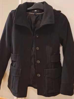 Svart kappa från H&M med fickor - Stilren svart kappa från H&M med stora knappar framtill, två rymliga fickor med lock och axelklaffar. Kappan har en klassisk krage och markerad midja med dekorativa knappar bak. Perfekt för kyliga dagar och enkel att matcha. St 36