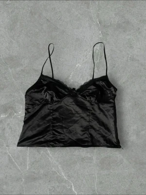 Svart satin linnetopp med spets - Stilren svart linnetopp i glansigt satinmaterial med tunna axelband och spetsdetaljer längs urringningen. Toppen har en liten rosett framtill och en mjuk, följsam passform som ger en elegant vibe. Perfekt att matcha med både jeans och kjol.