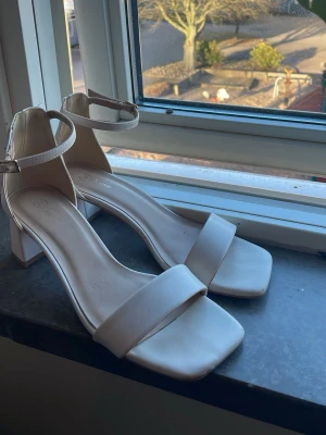 Vita klackar med rem - Snygga vita klackar med bred blockklack och fyrkantig tå. Skorna har en stilren rem runt ankeln med spänne och är tillverkade i ett slätt, matt material som ger en modern look. Perfekta för att lyfta vilken outfit som helst. Det står att de är storlek 37 men passar lite större så jag skulle säga 38. Har endast använts en gång!!