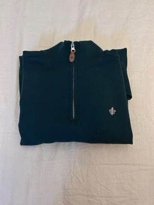 Mörkgrön halvzip-tröja Morris - Mörkgrön stickad tröja från Morris med halv dragkedja och ribbade muddar. Tröjan har en liten broderad logotyp på bröstet och kontrastfärgad insida på kragen. Perfekt för lager-på-lager och snygg till jeans.