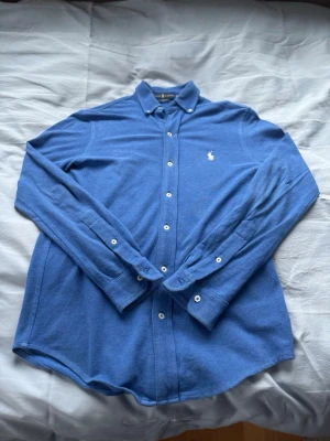 Blå skjorta från Ralph Lauren - Klassisk blå skjorta från Ralph Lauren med vit broderad logga på bröstet. Skjortan har lång ärm, knappar framtill och vid manschetterna samt en normal passform. Perfekt till både jeans och chinos för en clean look.