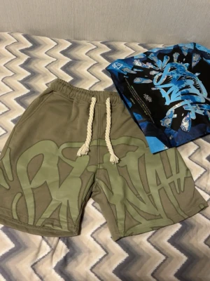 Olivgröna shorts med snörning - Säljer ett par olivgröna shorts med breda ben och coolt graffiti-inspirerat tryck i ljusare grönt. Shortsen har en tjock, vit dragsko i midjan och elastisk linning för skön passform. Perfekta för en avslappnad streetwear-look.