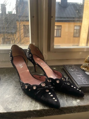 Svarta pumps med nitar från PRADA - Säljer ett par svarta pumps från PRADA med spetsig tå, dekorativa nitar och metallöglor längs kanterna. Skorna har en smal klack och är tillverkade i mocka. Behöver lite fix av en skomakare 