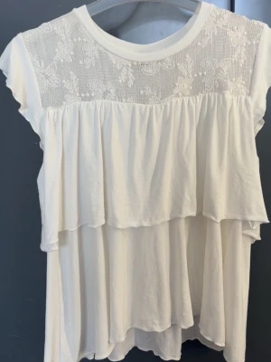 Vit blus med spets och volanger - Söt vit blus från kappahl med dubbla volanger och spetsdetaljer upptill. Blusen har korta ärmar och ett luftigt, flowy fall. Spetsen framtill har ett blommigt mönster. Storlek 158/164 men sitter som S