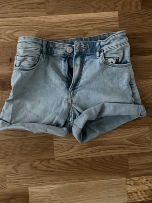 Ljusblå denimshorts med uppvik (vet inte varifrån) - Snygga ljusblå denimshorts med klassisk femficksmodell och uppvikta benslut. Shortsen har normal passform och stängs med knapp och dragkedja. Perfekta för varma dagar och enkla att matcha med olika toppar. Vet inte var de är ifrån.
